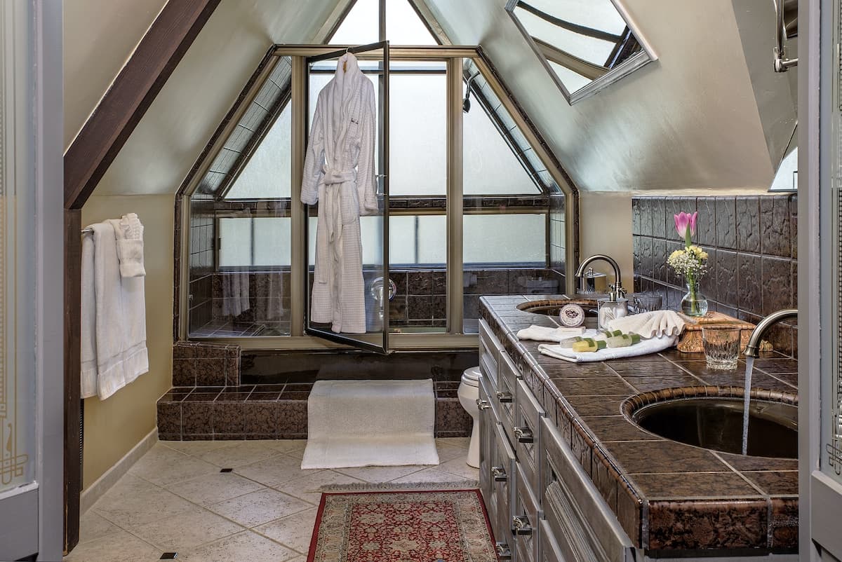 A modern bathroom with an angled ceiling features a glass shower, a marble countertop, a towel rack, and a robe hanging by the door.