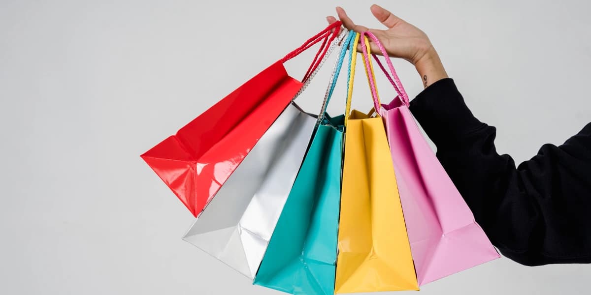 A hand holds five colorful shopping bags in various shades.