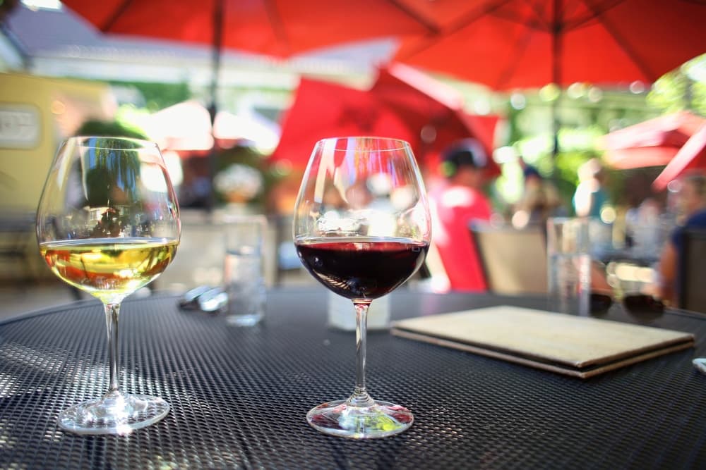 Two glasses of wine, one white and one red, on a black table surrounded by a lively outdoor setting.