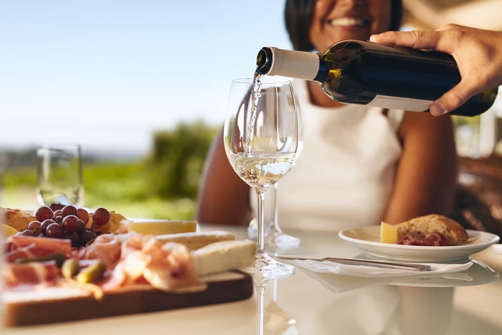 A person pours white wine into a glass at a table with a charcuterie platter and a smiling woman in the background.