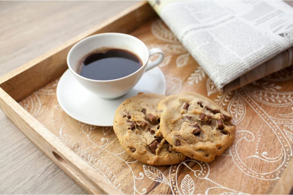 A cup of coffee with two chocolate chip cookies on a decorative wooden tray beside a rolled newspaper.