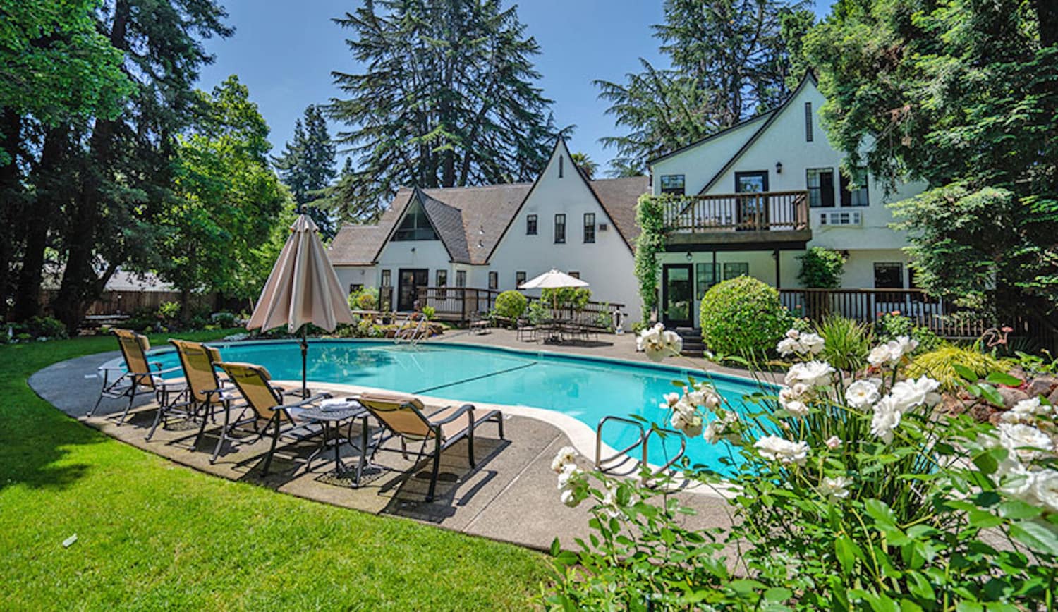 A serene backyard features a pool surrounded by lounge chairs, lush greenery, and a charming house in the background.