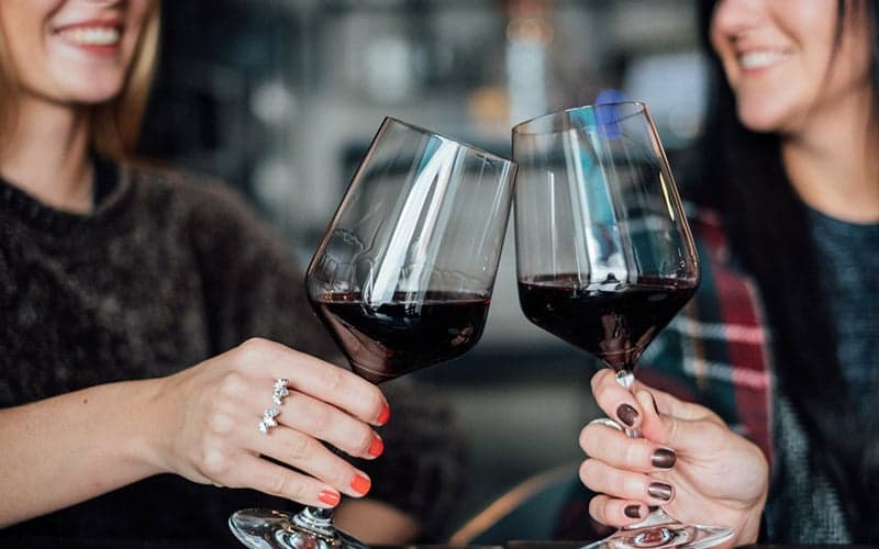 Two women clink glasses of red wine in a cozy setting.