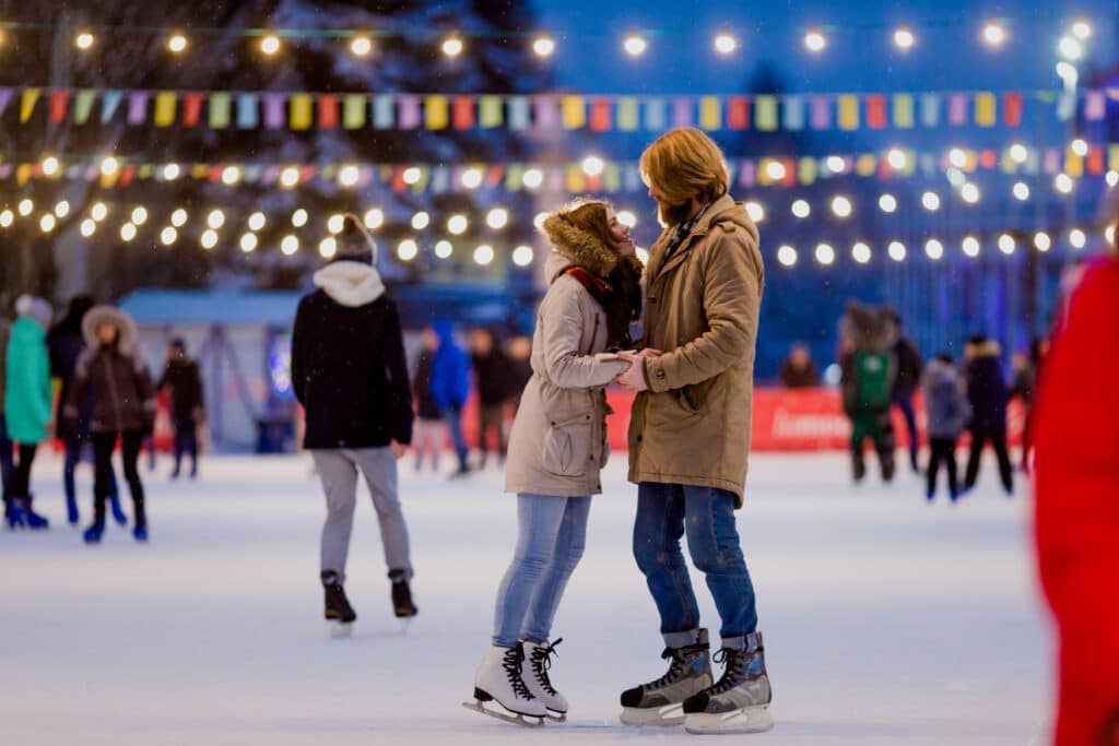 A couple skates close together on an ice rink, surrounded by festive lights and other skaters.