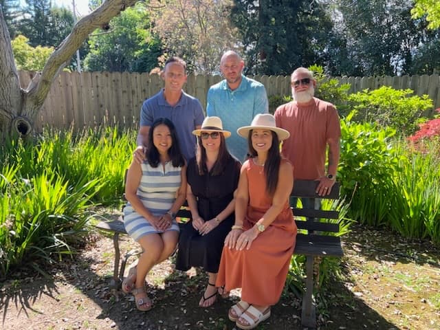A group of six people poses together on a bench in a sunny garden.