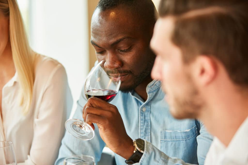 A man thoughtfully examines a glass of red wine while seated at a table with others.