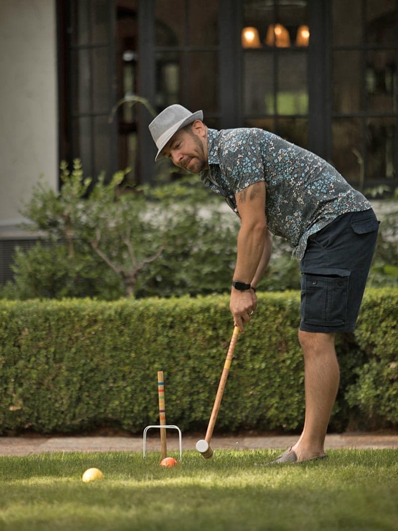 A man in a floral shirt and hat swings a croquet mallet on a grassy lawn.