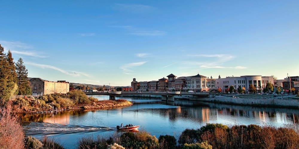 A scenic view of a river with a small boat and buildings lining the shore under a clear blue sky.