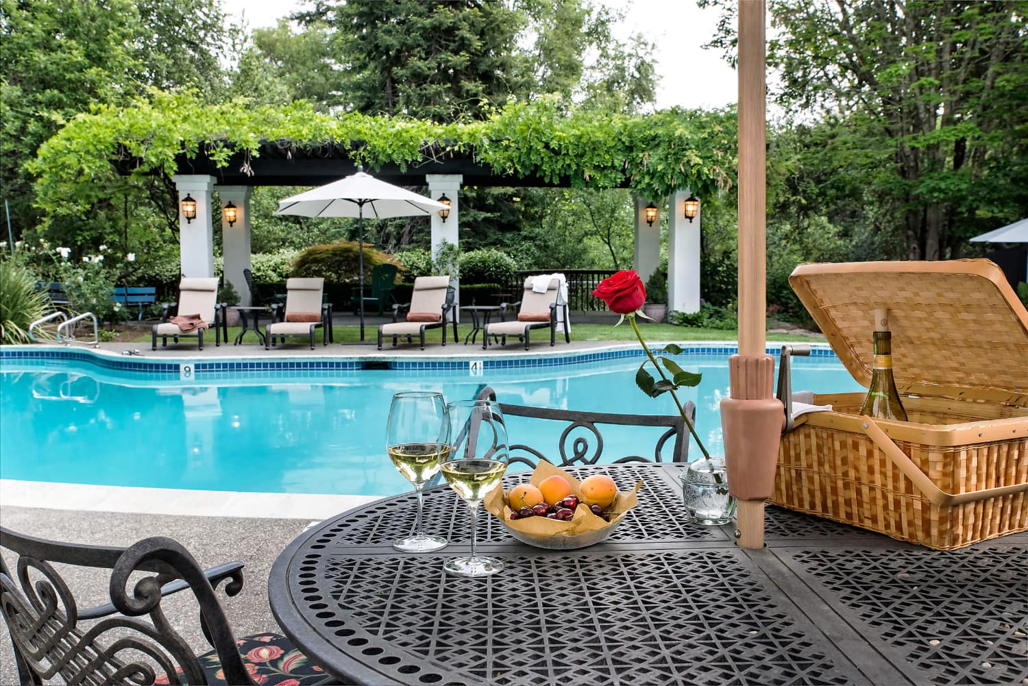 A table set for a picnic overlooks a swimming pool surrounded by lounge chairs and greenery.